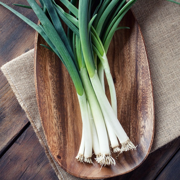 Dobies new vegetable seed 2024 Spring Onion 'Marksman' (Bunching Onion). Image shows 7 spring onions, cleaned and harvested, displayed on an oval wooden platter on a piece of hessian material, on top of a dark-wood table top.